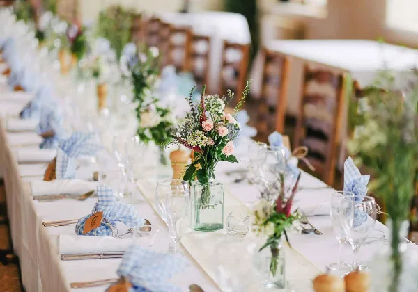A long wedding reception table set with white linens, blue gingham napkins, glassware, and floral centerpieces.