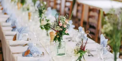 A long wedding reception table set with white linens, blue gingham napkins, glassware, and floral centerpieces.