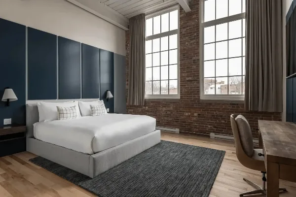 Interior of a boutique hotel room with a made bed, desk chair, wood floors, and large windows.