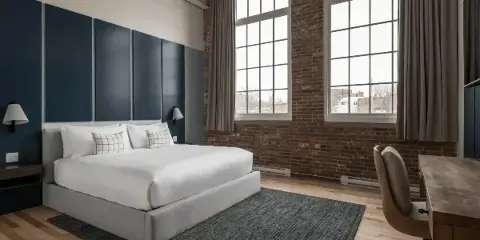 Interior of a boutique hotel room with a made bed, desk chair, wood floors, and large windows.