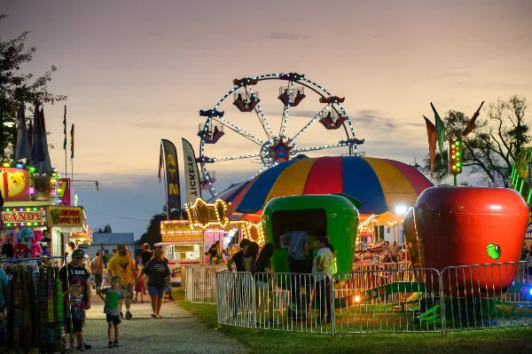 Evening view of the Miami County Fair midway featuring a Ferris wheel, colorful carnival rides, food booths, and visitors walking among the attractions.