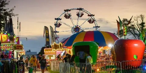 Evening view of the Miami County Fair midway featuring a Ferris wheel, colorful carnival rides, food booths, and visitors walking among the attractions.