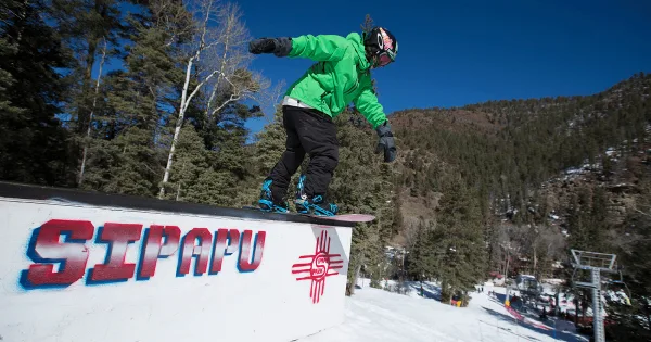A snowboarder in a green jacket performing a trick on a rail with the Sipapu logo, set against a snowy mountain landscape.