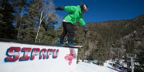 A snowboarder in a green jacket performing a trick on a rail with the Sipapu logo, set against a snowy mountain landscape.