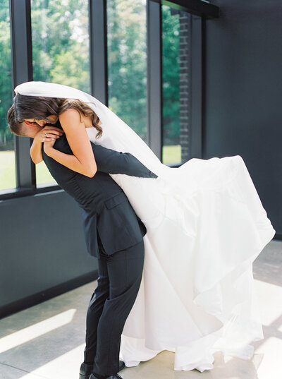 A bride in a white gown and veil embracing a groom in a black suit near a large window.