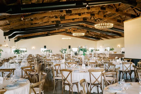 Interior view of a wedding reception hall with round tables, wooden chairs, and exposed timber ceiling beams.