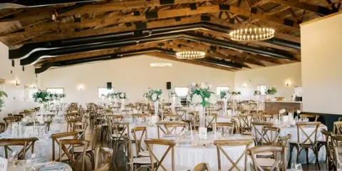 Interior view of a wedding reception hall with round tables, wooden chairs, and exposed timber ceiling beams.