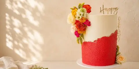 A two-tiered cake with red textured fondant and white fondant, decorated with a cluster of colorful fresh flowers and a gold 'Happy Birthday' topper, sitting on a white cake stand.