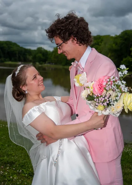 A bride in a white gown and a groom in a pink suit embracing outdoors near a body of water.