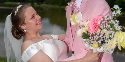 A bride in a white gown and a groom in a pink suit embracing outdoors near a body of water.