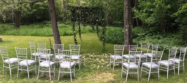 An outdoor wedding ceremony setup with white Chiavari chairs arranged on a lawn facing a floral arch.