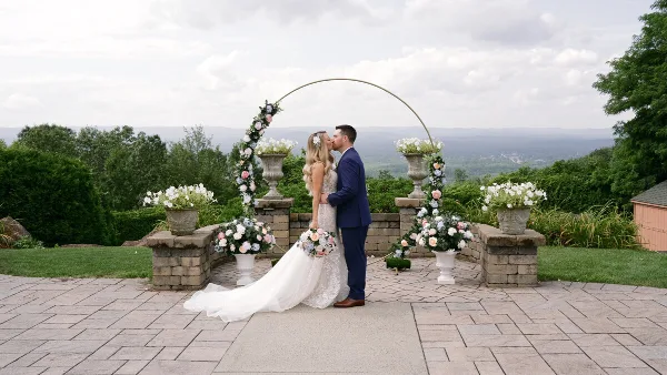 A bride and groom sharing a kiss at a wedding ceremony on an outdoor stone patio with floral decorations and a scenic background.