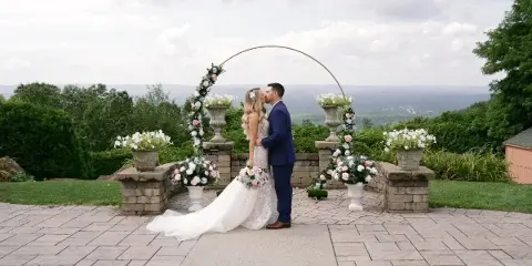 A bride and groom sharing a kiss at a wedding ceremony on an outdoor stone patio with floral decorations and a scenic background.