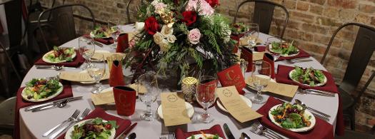 A round table set with plated food, red napkins, and a large floral centerpiece in a venue with a brick wall.
