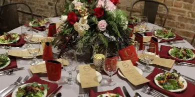 A round table set with plated food, red napkins, and a large floral centerpiece in a venue with a brick wall.