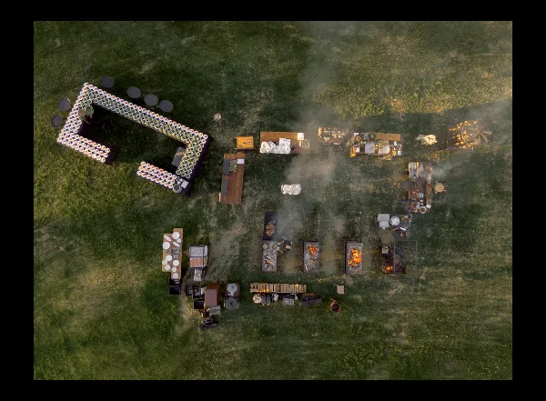 Aerial view of an outdoor catering event with open-fire cooking stations and food tables on a grassy field.