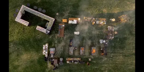 Aerial view of an outdoor catering event with open-fire cooking stations and food tables on a grassy field.