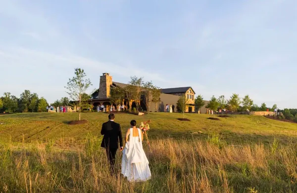 A bride and groom walking through a grassy field toward the Bennett Farms venue building.