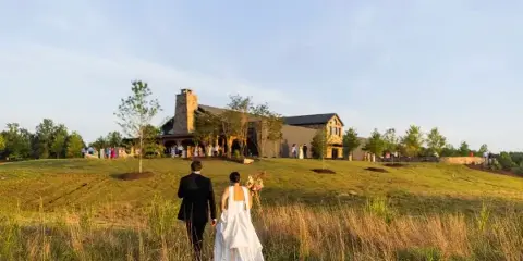 A bride and groom walking through a grassy field toward the Bennett Farms venue building.
