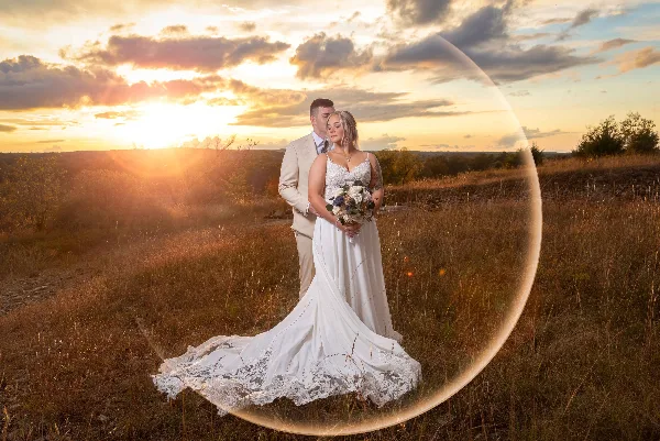 A bride and groom standing in a grassy field at sunset, with the bride holding a bouquet and a large lens flare visible in the foreground.