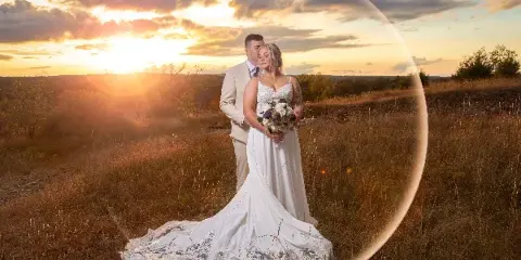 A bride and groom standing in a grassy field at sunset, with the bride holding a bouquet and a large lens flare visible in the foreground.