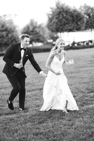 A black and white photo of a bride and groom running through a grassy field, holding hands and drinks.