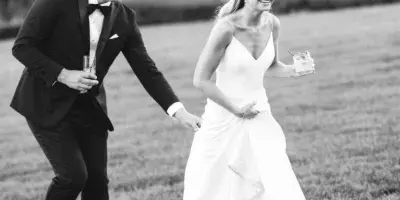 A black and white photo of a bride and groom running through a grassy field, holding hands and drinks.