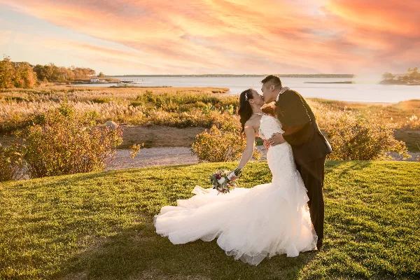 A bride and groom sharing a kiss on a grassy hill overlooking a body of water during a sunset.
