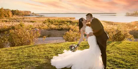 A bride and groom sharing a kiss on a grassy hill overlooking a body of water during a sunset.
