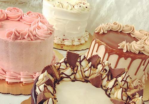 A display of various decorated cakes, including a pink frosted cake, a white tiered cake, and a chocolate cake with chocolate shavings.