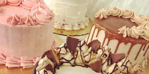 A display of various decorated cakes, including a pink frosted cake, a white tiered cake, and a chocolate cake with chocolate shavings.