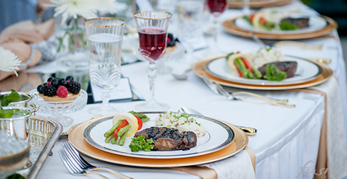 A formal dining table set with white linens, featuring plated meals with meat and vegetables, wine glasses, and floral centerpieces.
