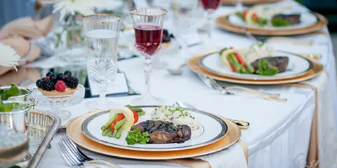 A formal dining table set with white linens, featuring plated meals with meat and vegetables, wine glasses, and floral centerpieces.