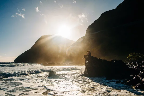 A couple sitting on a rocky outcrop overlooking the ocean at sunset, silhouetted against a bright sky.