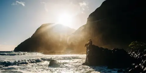 A couple sitting on a rocky outcrop overlooking the ocean at sunset, silhouetted against a bright sky.