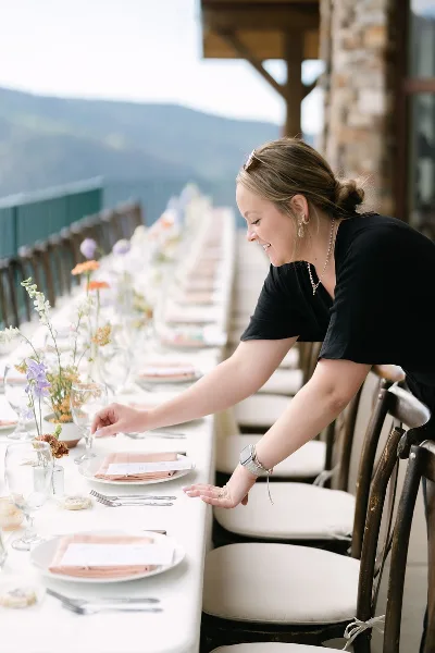 A wedding planner adjusting a table setting with floral centerpieces and place settings at an outdoor venue.