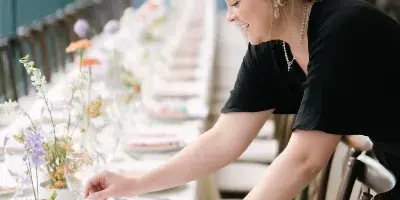 A wedding planner adjusting a table setting with floral centerpieces and place settings at an outdoor venue.