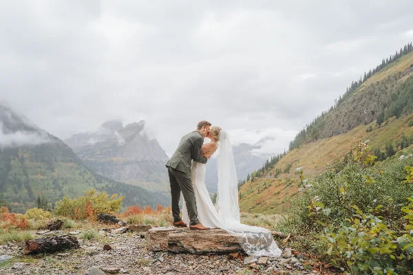 A bride and groom share a kiss on a wooden log in a mountainous landscape with misty peaks in the background.