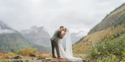 A bride and groom share a kiss on a wooden log in a mountainous landscape with misty peaks in the background.
