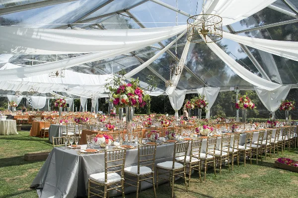 An outdoor wedding reception setup under a clear tent with white drapery, gold chairs, and floral centerpieces.