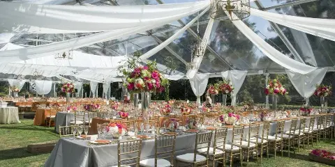 An outdoor wedding reception setup under a clear tent with white drapery, gold chairs, and floral centerpieces.