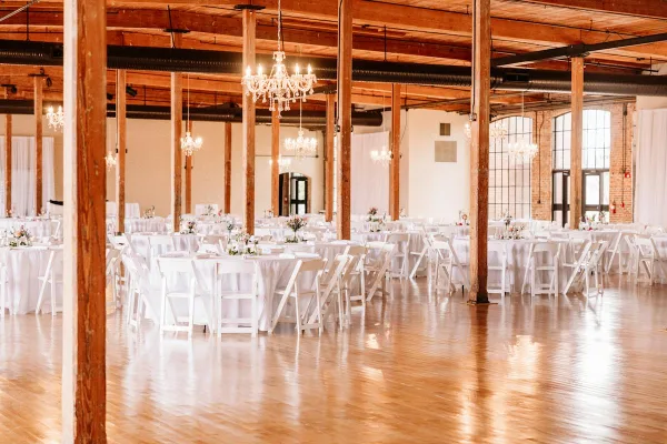 Interior view of a large event hall with wooden pillars, chandeliers, and round tables set for a wedding reception.