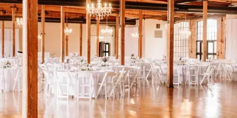 Interior view of a large event hall with wooden pillars, chandeliers, and round tables set for a wedding reception.
