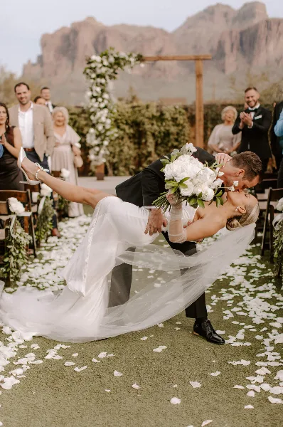 A bride and groom share a kiss during an outdoor wedding ceremony with a floral arch and guests in the background.