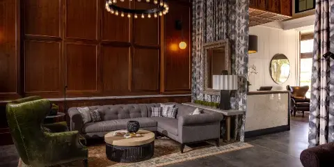 Interior view of The Louis Hotel lobby featuring a grey sectional sofa, green armchair, wood paneling, and a chandelier.