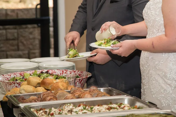 A bride and groom serving themselves salad and other dishes from a catering buffet.