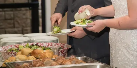 A bride and groom serving themselves salad and other dishes from a catering buffet.