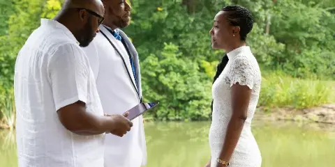 A bride and groom standing outdoors near a body of water, with the groom holding a tablet.