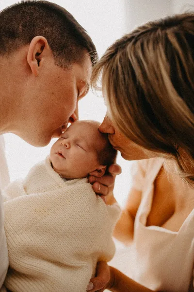 A close-up photograph of a man and a woman kissing the forehead of a sleeping newborn baby wrapped in a white knit blanket.