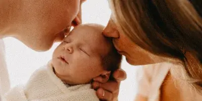 A close-up photograph of a man and a woman kissing the forehead of a sleeping newborn baby wrapped in a white knit blanket.